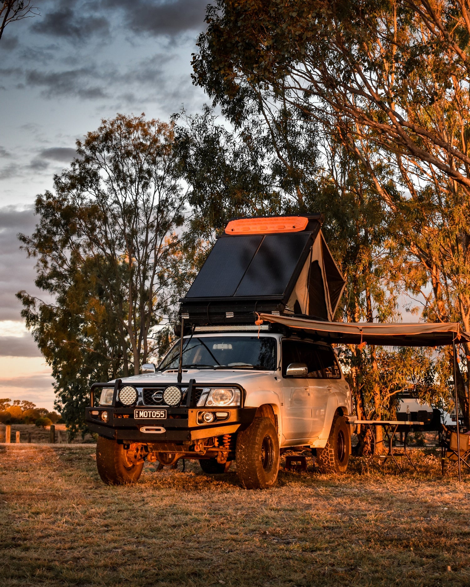 White Nissan Patrol with a rooftop tent parked in a natural setting with trees and a cloudy sky.